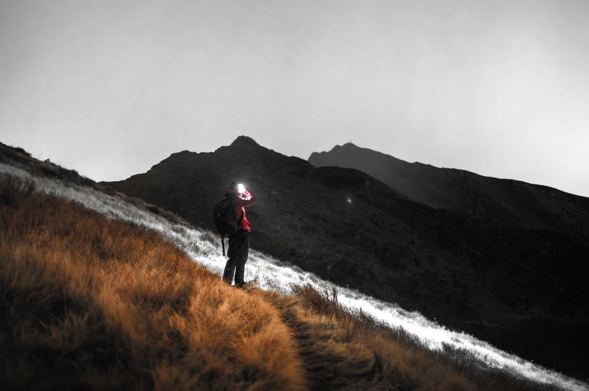A lone hiker illuminated by a headlamp traverses a mountain path under a cloudy sky.